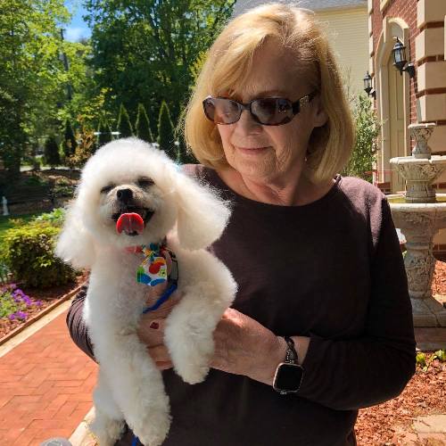 Smiling woman holds a fluffy white dog with its tongue out, wearing a colorful patterned bandana. Sunny outdoor setting with a brick walkway and a fountain in the background.