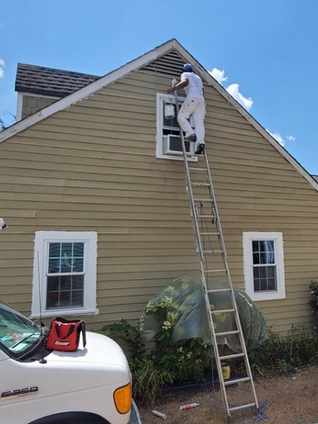 A painter on a ladder works on exterior house painting, reaching a window above an air conditioner. The house is light brown with white trim, under a sunny blue sky.