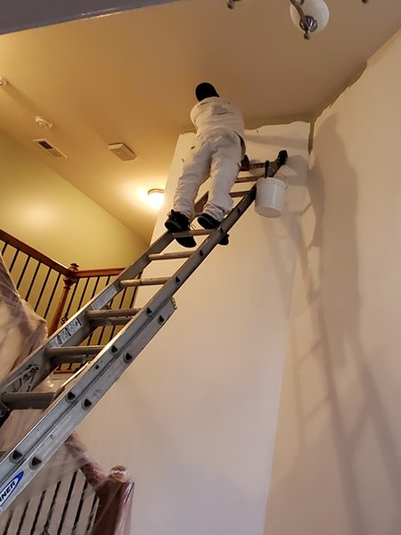 A painter in white coveralls stands on a ladder, carefully painting the upper corner of an interior wall during an interior home painting project. A paint bucket hangs from the ladder.